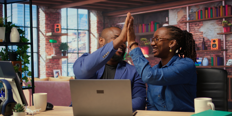 Black freelancing couple doing a high five to celebrate their achievement, completing tasks to ensure successful project. Young man and woman feeling pleased with their cooperation. Camera A.