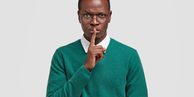 Close your mouth and shut up please. Dark skinned serious male model keeps index finger over mouth, asks not spread rumors, wears spectacles and green jumper, isolated over white background.